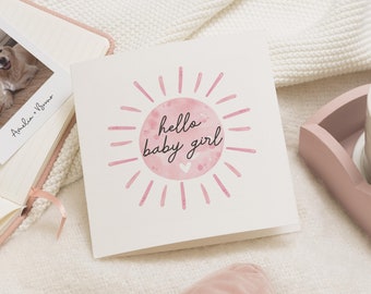 May include: A square greeting card with the text "hello baby girl" in black script, surrounded by a pink sunburst design. The card is on a white surface, with a photo album and a pink tray in the background.