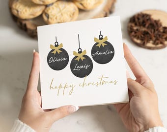 May include: A white Christmas card with a gold inscription that reads "happy christmas". Three black ornaments with gold bows hang from the top of the card. Each ornament has a name written on it: Olivia, Lewis, and Amelia.