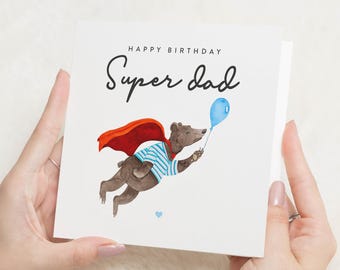 May include: A birthday card with a cartoon bear wearing a red cape and blue and white striped shirt, holding a blue balloon. The card says "Happy Birthday Super Dad".