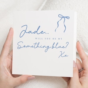 May include: A square, off-white card with blue script that reads "Jade... Will you be my Something blue?" and a blue bow illustration. The card is held by hands with a ring on one finger.