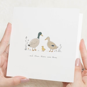 Op de afbeelding: Een vierkante witte wenskaart met een centrale illustratie van twee volwassen eenden en een klein geel eendenkuiken in het gras met wilde bloemen. Onder de eenden staat de tekst: "...and then there were three."