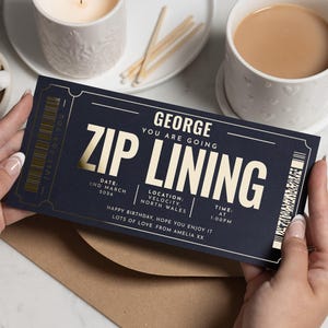 May include: A navy blue invitation card with gold lettering that reads "ZIP LINING" and "GEORGE YOU ARE GOING". The card includes the date, location, and time. The card is held by two hands, with a cup of coffee and a lit candle in the background.