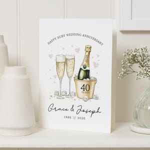 May include: A white greeting card featuring a watercolor illustration of two champagne flutes and a bottle in a gold ice bucket. The text reads Happy Ruby Wedding Anniversary, 40 Years, and the names Grace & Joseph with the years 1986 and 2026.