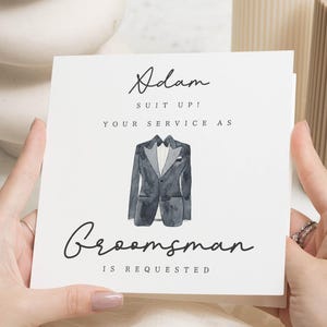 Op de afbeelding: Een vierkante witte kaart met zwarte tekst: "Adam suit up! Your service as Groomsman is requested". Een aquarel illustratie van een zwart smokingjasje staat centraal op de kaart. De kaart wordt door twee handen vastgehouden.