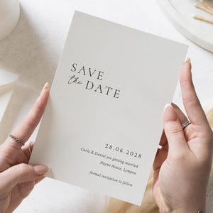 Op de afbeelding: Een crèmekleurige Save the Date-kaart met zwarte tekst. De kaart luidt "SAVE the DATE" in een scriptlettertype, gevolgd door de datum, namen en locatie. De kaart wordt door twee handen vastgehouden.