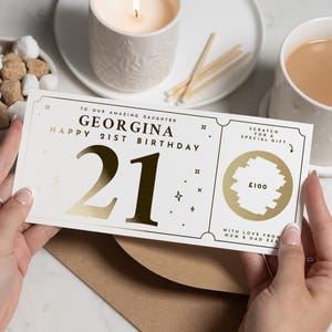 Peut inclure: Une carte à gratter personnalisée pour un 21e anniversaire, avec le texte "Happy 21st Birthday Georgina" en or. La carte présente une zone à gratter dorée avec "£100" imprimé en dessous. Une bougie allumée, une tasse de thé et des sucres sont en arrière-plan.