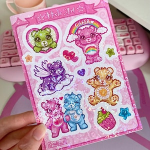 May include: A pink sticker sheet with holographic accents featuring various cartoon bears with different expressions. The sheet includes the text "Hxlx Art" and "Cheer".