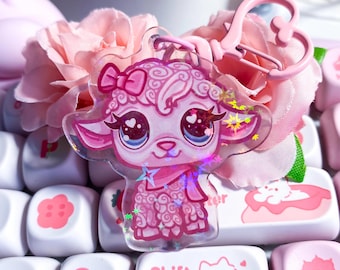 Op de afbeelding: Een roze acryl sleutelhanger met een cartoon schaap met hartvormige ogen, een strik en een sjaal. De sleutelhanger is bevestigd aan een roze hartvormige sluiting. De achtergrond omvat roze bloemen en een toetsenbord met roze toetsen.