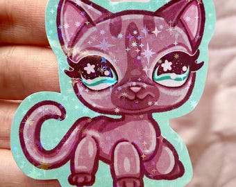 May include: A pink and purple glitter cat sticker with sparkling white stars and large blue eyes. The cat is sitting on a light blue background.