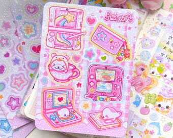 May include: A sheet of pink stickers with a variety of cute illustrations. Designs include handheld gaming consoles, a bear in a teacup, and a seal on a book. The stickers have a gingham pattern background and the text "Halo Art" is visible.