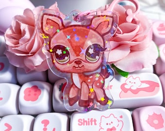 May include: A pink deer-shaped keychain with a holographic finish, featuring star accents. The keychain has a pink clasp and is set against a backdrop of pink roses and white keyboard keys with pink illustrations.