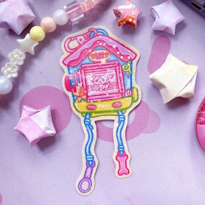 May include: A vibrant sticker of a cartoon house with a panda inside. The house is pink, blue, and yellow, with a dog bone and a microphone attached. Origami stars and a beaded bracelet surround the sticker on a pink surface.