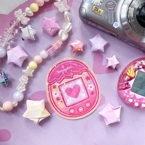 May include: A collection of pink and white origami stars, a beaded necklace, and a pink Tamagotchi sticker with a heart design. A digital camera and a pink Tamagotchi device are also present, all on a lavender surface.