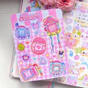 Könnte beinhalten: Ein Bogen mit Aufklebern mit farbenfrohen Illustrationen von Retro-Handheld-Elektronikspielen und niedlichen Charakteren. Die Aufkleber haben einen rosa-weiß karierten Hintergrund und den Text "Hello Art". Weitere Aufkleber sind Herzen, Sterne und ein Hase.