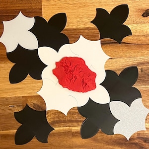 May include: A decorative floral design composed of black, white, and silver petal-shaped tiles. The center features a textured red element. The tiles are arranged on a wooden surface, creating a striking contrast of colors and textures.