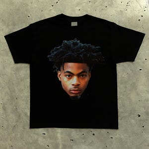May include: Black t-shirt featuring a detailed portrait of a man with dark, curly hair. The portrait is centered on the shirt and displays the man's face and upper chest. The shirt is made of a soft material.