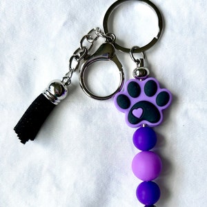May include: Purple and black paw print keychain with a small heart on the paw print. The keychain has a silver ring and a black tassel.