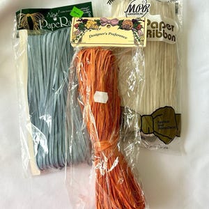 May include: Three packages of paper ribbon in light blue, bright orange, and off-white. Each bundle is wrapped in clear plastic packaging with branded labels. The labels feature text including PaperRaffia, Designer's Preference, and Paper Ribbon.