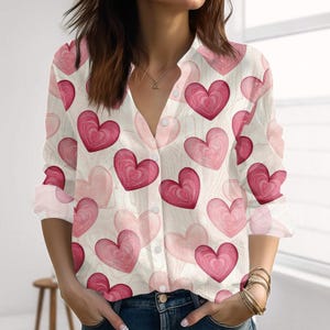 Puede incluir: Camisa abotonada blanca adornada con un estampado de corazones rosas y rojos. La camisa de manga larga tiene cuello en V y está hecha de un material ligero. Los corazones varían en tamaño y tono, creando un diseño romántico y femenino.