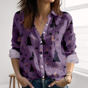 Puede incluir: Camisa morada con botones con estampado de brujas de Halloween. La camisa de manga larga presenta un patrón repetido de figuras de brujas negras, calabazas y elementos florales. La camisa tiene cuello y puños abotonados.
