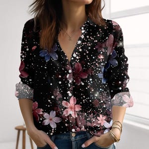 Könnte beinhalten: Schwarzes Langarmhemd mit floralem Muster in Rosa, Lila und Blau. Das Hemd hat einen V-Ausschnitt und weiße Sprenkel. Die Ärmel sind hochgekrempelt und zeigen eine graue Manschette. Das Hemd ist vorne geknöpft.