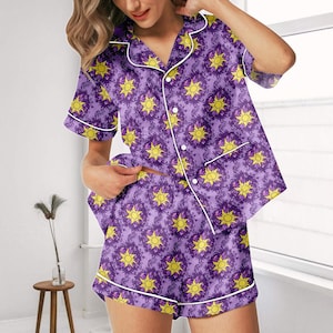 Könnte beinhalten: Lila Pyjama-Set mit einem sich wiederholenden gelben Sonnenmuster. Das kurzärmelige Oberteil hat weiße Paspeln und eine Knopfleiste. Die passenden Shorts haben ebenfalls weiße Paspeln. Das Set ist aus einem weichen, leichten Stoff gefertigt.