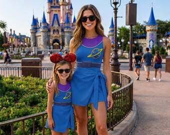 May include: Two matching blue sleeveless dresses with purple necklines, a waist belt detail, and a yellow planet graphic on the chest. The dresses feature a wrap-style skirt with a side tie. The child wears a red sequined mouse ear headband.
