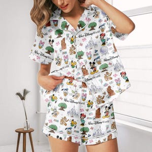 May include: White pajama set with a Disney theme, featuring characters, castles, and trees. The set includes a short-sleeved button-up top and matching shorts. The text "Walt Disney World" and "2026" are printed on the fabric.