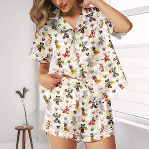 Peut inclure: Un ensemble de pyjama blanc composé d'une chemise à manches courtes boutonnée et d'un short assorti, orné d'un motif répétitif de personnages de dessins animés déguisés pour Halloween, incluant des fantômes, des diables et des citrouilles.