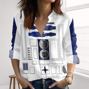 Könnte beinhalten: Weißes Button-Down-Shirt mit blauem und schwarzem Grafikdesign. Das langärmlige Shirt hat ein geometrisches Muster, das einem Roboter ähnelt. Die Ärmel und Seiten des Shirts sind blau.