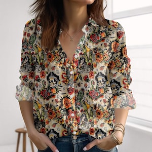 Könnte beinhalten: Ein langärmliges Button-Down-Shirt mit einem lebendigen Allover-Print mit Totenköpfen, Rosen, Kakteen und weiblichen Figuren. Das Shirt hat einen V-Ausschnitt und ist in Beige-, Rot-, Orange- und Grüntönen gehalten.