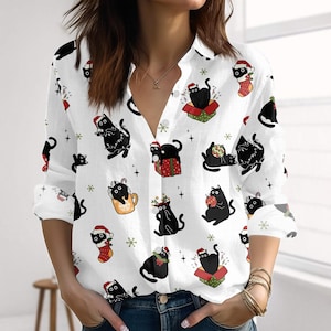 Könnte beinhalten: Weißes Hemd mit einem sich wiederholenden Muster aus schwarzen Katzen in festlicher Kleidung. Die Katzen tragen Weihnachtsmützen, sitzen in Geschenkboxen und halten Ornamente. Das Hemd hat lange Ärmel und einen V-Ausschnitt.