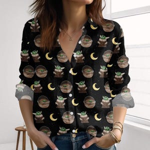 Könnte beinhalten: Schwarzes Button-Down-Shirt mit einem sich wiederholenden Muster eines grünen Alien-Charakters in einer braunen Trage und gelben Halbmonden. Das langärmlige Hemd hat einen V-Ausschnitt und ist aus einem weichen Material gefertigt.