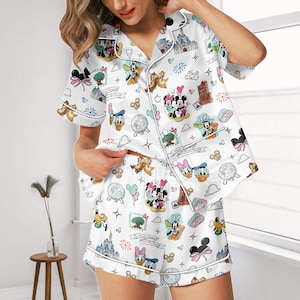 May include: White Disney-themed pajama set. The short-sleeved top and shorts are decorated with Disney characters, castles, and other graphics. The set has white piping and a button-down top. The measurements are in centimetres.