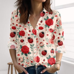May include: A cream-coloured button-down shirt featuring a repeating pattern of red roses, pink flowers, green leaves, and ladybirds. The long-sleeved shirt has a V-neck and is made of a lightweight fabric.