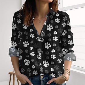 May include: Black button-down shirt featuring a repeating pattern of white and grey paw prints. The long-sleeved shirt has a V-neck and rolled-up sleeves. The shirt is made of a soft, lightweight material.