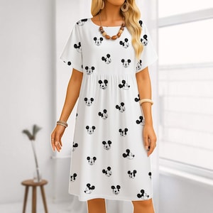 Könnte beinhalten: Weißes, kurzärmeliges Kleid mit einem Muster aus schwarzen Mickey Mouse-Gesichtern. Das Kleid hat einen runden Ausschnitt und eine geraffte Taille. Das Model trägt Schmuck, darunter eine Halskette und Armbänder. Das Kleid ist knielang.