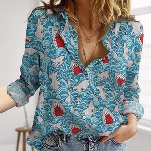 May include: A light blue button-down shirt featuring a repeating pattern of sharks with red mouths and white teeth set against a backdrop of blue waves. The shirt has long sleeves and a collared neckline. The fabric appears lightweight.