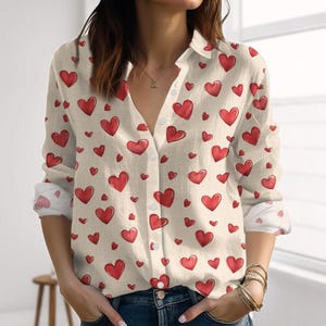 Op de afbeelding: Crèmekleurig overhemd met knopen en een patroon van rode harten. Het shirt met lange mouwen heeft een kraag en is aan de voorkant dichtgeknoopt. De manchetten van de mouwen zijn wit met een rood hartpatroon.