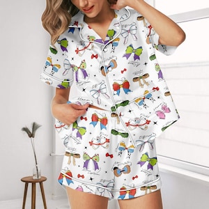 May include: White satin pajama set with a colorful bow pattern. The short-sleeved top and shorts feature various bow designs in red, blue, green, and purple, with black text accents. The set has white piping and a button-down front.