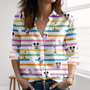 Puede incluir: Camisa blanca con botones y un patrón de rayas coloridas. La camisa presenta varios personajes de dibujos animados, incluyendo Mickey Mouse, Minnie Mouse, Donald Duck y Goofy. Las rayas son en tonos rosa, naranja, amarillo, verde, morado y azul.