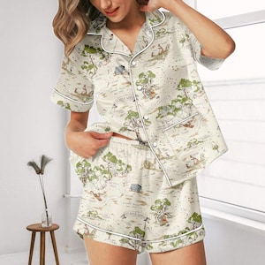 Pode incluir: Conjunto de pijama creme com um design semelhante a um mapa com personagens de desenhos animados e árvores. O conjunto inclui uma camisa de manga curta com botões e calções a condizer, ambos com debrum branco.
