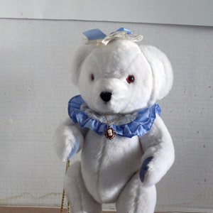 May include: White teddy bear wearing a blue and white floral collar with a cameo pendant. The bear has red eyes and blue feet. The bear is sitting on a gold stand.