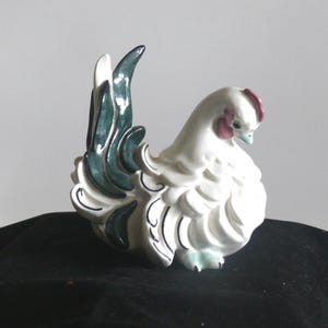 May include: White ceramic chicken figurine with green and black painted details. The chicken has a raised tail and a pink comb.
