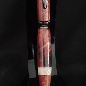 May include: A wooden pen with a red and white wood grain pattern. The pen has a silver metal band and a black tip. The pen is sitting on a black surface. P24