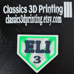 May include: A white baseball-shaped plaque with a green border and green leaf accents. The name "ELI" is printed in navy blue letters and the number "3" is printed in navy blue below the name.