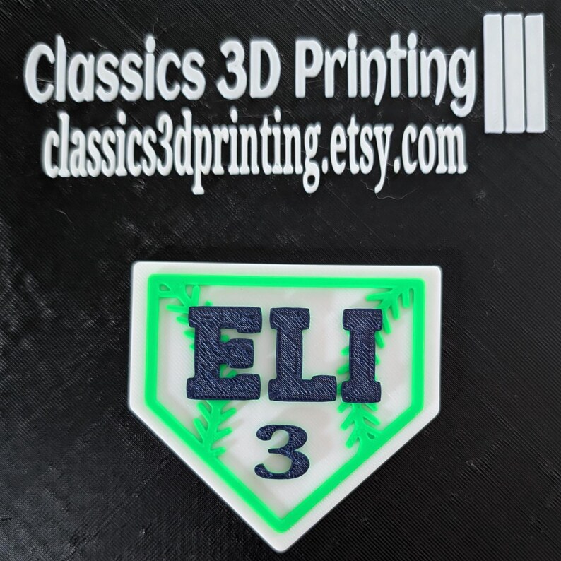 May include: A white baseball-shaped plaque with a green border and green leaf accents. The name "ELI" is printed in navy blue letters and the number "3" is printed in navy blue below the name.