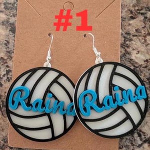 May include: A pair of white volleyball earrings with black lines and the name "Raina" in blue lettering. The earrings are on a brown card with the number "#1" written in red.