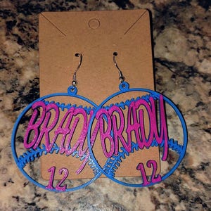 May include: A pair of blue and pink baseball-themed earrings with the name "BRADY" and the number "12" on each earring.
