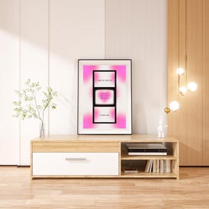May include: A framed print with a pink gradient background and a black and white image of a television screen with the text "Verse of the Day" and a Bible verse. The print is hanging on a wall above a wooden TV stand.